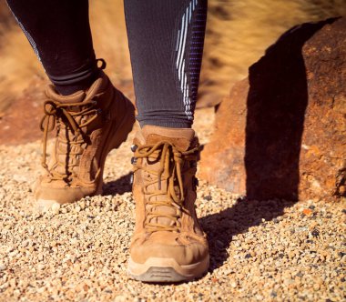Uzun bacaklı çakal çizmeleri ve desenli taban tabanı olan bir kız, dağlarda bir patika boyunca yürüyor. Aktif bir eğlence için rahat ayakkabılar giymiş kayalık bir arazi..