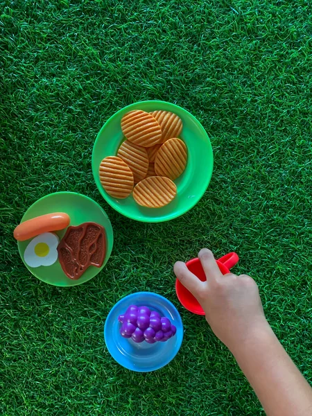 Plates with food and beverages toys on grass. Children\'s hands play cooking.