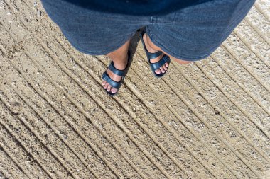 Beyaz kumların üzerinde duran siyah sandaletli bir çift ayak.