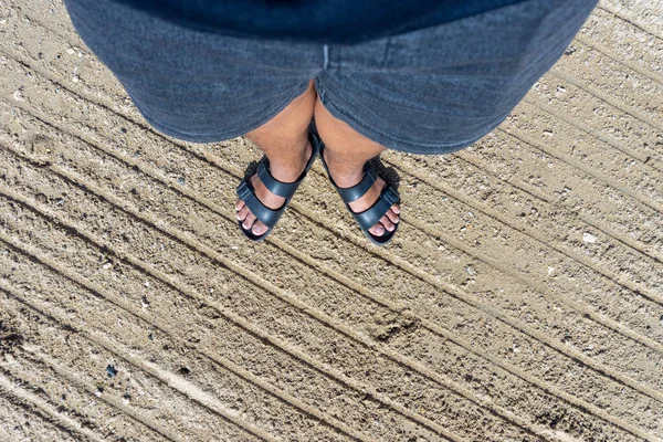 A pair of feet in black sandals standing on white sand