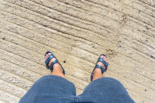 A pair of feet in black sandals standing on white sand
