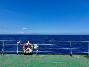 Feribot güvertesinde demir parmaklıklar ve açık deniz ve mavi gökyüzüne bakan can simidi.