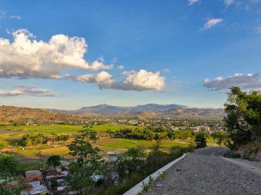 Yeşil pirinç tarlaları, dağınık evler ve öğlen güneşinin altındaki uzak dağlarla dolu tepeden Bima şehrinin çarpıcı manzarası.