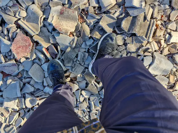 Kırık kiremitler ve öğlen güneşinin altında beton döküntülerin üzerinde duran siyah beyaz spor ayakkabılarla bir adamın ayaklarının yakın çekimi..