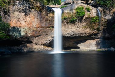 Long exposure effect of flowing water of rock cliff waterfall on sunny day inside tropical rainforest.
