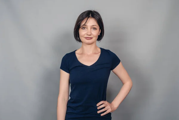 Beautiful woman with dark hair wearing  dark t-shirt. Looking at  camera. Gray background .Hand on hip.
