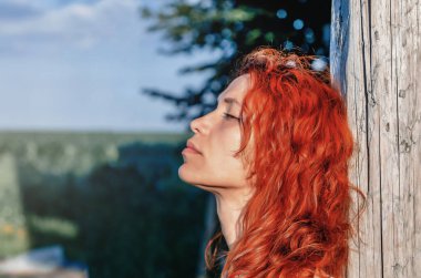 Red-haired woman in profile on the nature. Pressed against wooden pole