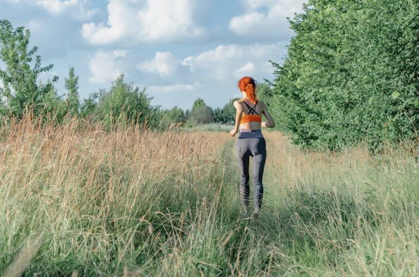 A young female runner runs along the road overgrown with grass. Rear view