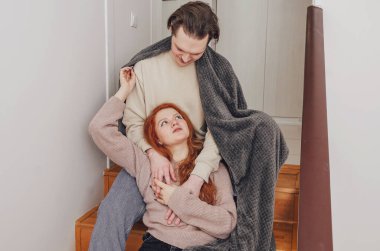 Guy, wrapped in blanket, gently hugs girl, looking at her. The girl dreamily sits on arms of her lover.