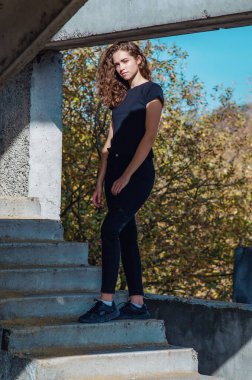 Curly-haired woman in black jeans stands on stairs. Abandoned building.