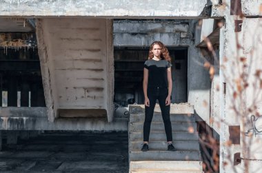 Curly-haired woman in black jeans stands on stairs. Abandoned building.