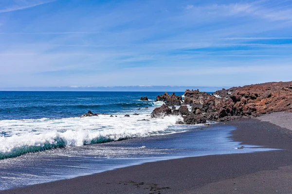 stock image Bright contrast of white sea foam of the waves rolling onto shore with the sand and black solidified lava.