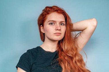 Siyah tişörtlü ciddi kızıl saçlı kadın gri arka planda duruyor. Eller saçın yanında.
