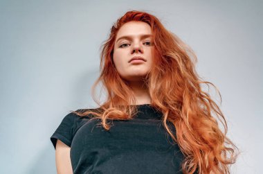 Siyah tişörtlü ciddi kızıl saçlı kadın gri arka planda duruyor. Aşağıdan görüntüle