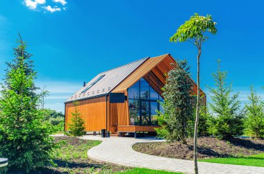 Mavi gökyüzünün altında patikası, ağaçları ve çalıları olan küçük bir ahır evi.