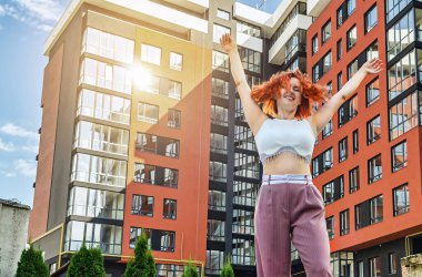 Neşeli kızıl saçlı kadın ellerini kaldırdı, yeni modern evin arka planında sevinçle