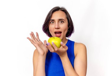 Elinde yeşil elma tutan heyecanlı bir kadın. Kameraya bakıyor. Mavi elbise, beyaz arka plan.