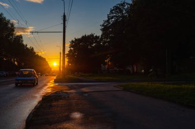 Şehirde gün batımlı asfalt yol. Yol boyunca park edilmiş araba sayısı. Fenerler ve ağaçlar. Alacakaranlık