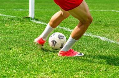 Genç futbolcu yeşil bir sahada iş başında görülüyor. Kırmızı şort giyen oyuncu futbol topunu kontrol ediyor..