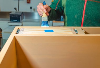 Marangoz, marangozluk atölyesinde fırçayla bir tahtanın yüzeyine yapıştırıcı sürer.