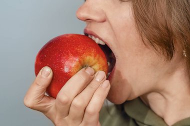 Kadının yüzünün bir parçası kırmızı elmayı ısırıyor, dişlerini gösteriyor. Sağlıklı yiyecekler. Gri arkaplanda yakın plan görünümü