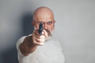 Gözlüklü kel sakallı adamın resmi. Kameraya silah doğrultuyor. Gri arka plan. Tehlike, tehdit
