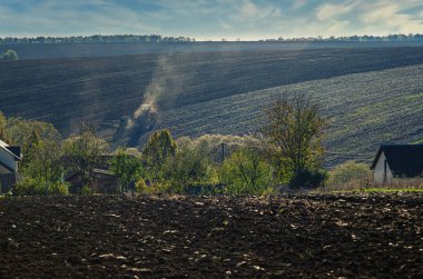 Traktör tarlası. Köyün ağaçlarının ve evlerinin arasından tarlaya bakın. Fırtınalı gökyüzü
