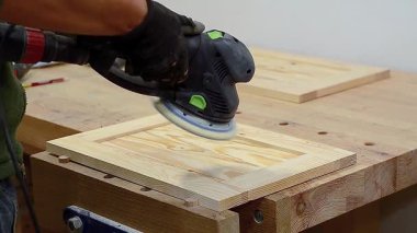 Marangoz, bir marangoz atölyesinde elektrikli zımpara makinesi kullanarak ahşap bir kapıyı zımparalıyor.