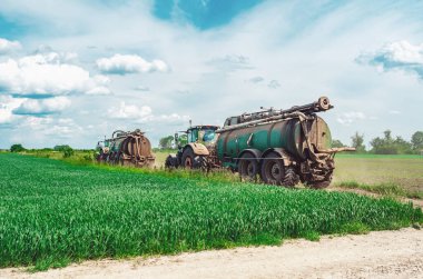 Organik gübre ve gübre dolu tankları olan çiftlik traktörlerinin yan görüntüsü. Yeşil alanlar arasında giden traktörler