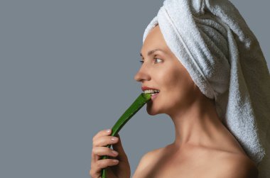 Gri arka planda, elinde aloe vera filizi tutan ve beyaz dişleriyle ısıran mutlu kadının yan görüntüsü. Yaşlanma karşıtı bakım konsepti. Boşluğu kopyala.