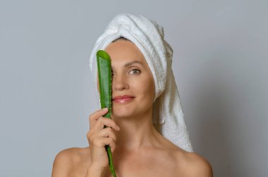 Beyaz havluya sarılı beyaz bir kadın bir gözünü aloe yaprağı ile kaplıyor ve gülümsüyor. Gri stüdyo geçmişi. Kaplıca ve cilt bakımı.