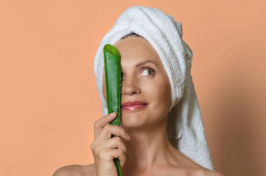 Beyaz havluya sarılı beyaz bir kadın bir gözünü aloe yaprağı ile kaplıyor ve gülümsüyor. Şeftali rengi stüdyo. Kaplıca ve cilt bakımı.