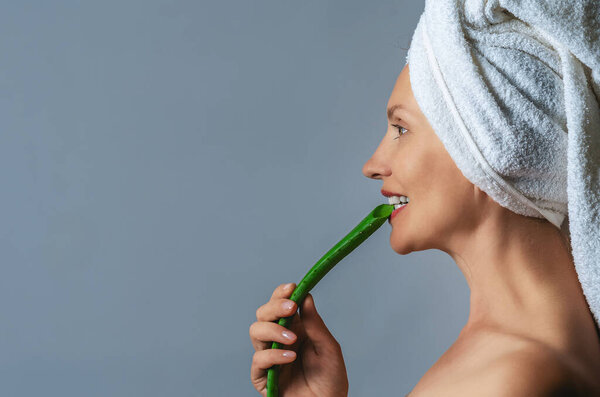 Вид сбоку счастливой женщины на сером фоне, держащей в руке веточку алоэ вера и кусающей ее белыми зубами. Концепция антивозрастного ухода. Копирование пространства.