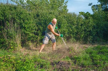 Sakallı, kel, gözlüklü, pantolonlu ve tişörtlü güçlü bir adam otları tırpanla biçiyor. Ev bakımı.