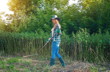 Şapkalı, ekose gömlekli, kot pantolonlu ve lastik çizmeli, güneşli bir günde otları tırpanla biçen sırıtan sarışın bir kadın. Ev bakımı.