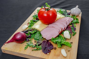 Dilimlenmiş jambon, peynir ve sebze. Dilimlenmiş peynirli, jambonlu, domatesli, biberli, sarımsaklı ve otlu restoran panosu. Sağlıklı protein menüsü. Siyah arkaplan.