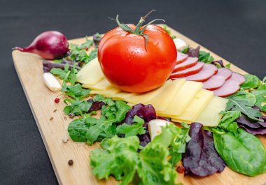 Jambon, peynir ve sebze dilimleri. Dilimlenmiş peynirli, jambonlu, domatesli ve biberli restoran panosu, salata ve yeşillikle servis ediliyor. Sağlıklı protein menüsü. Siyah arkaplan.