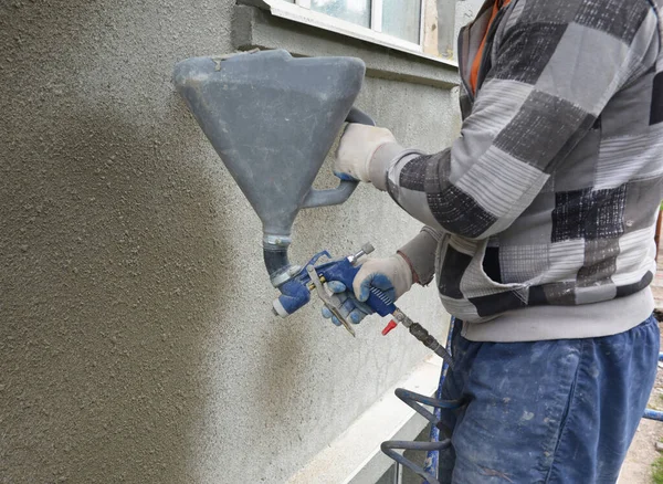 A builder is rendering, plastering, coating house  exterior wall of a building using plaster sprayer machine, stucco concrete sprayer gun.
