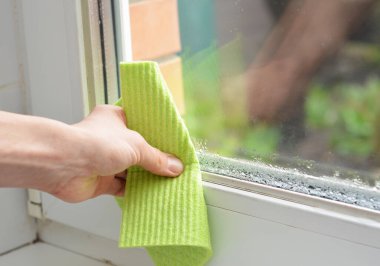 Bir kadın, yüksek nem seviyesiyle odadaki su buharını temizler. Pencere camında su birikintisi olan ev nemi..