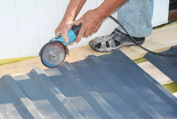 A building contractor is cutting lightweight metal, steel, corrugated, paint coated roofing sheet with an electric angle grinder laying profile sheet on the ground across padded support.