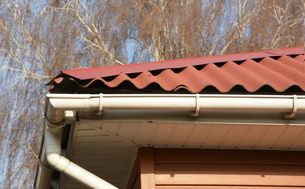 Close up on house roof corner with rain gutter pipeline, fascia board and wave profiled bituminous sheets.