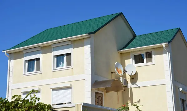 A modern detached house with a green metal roof, stucco, plastered painted walls, windows with closed plastic rolling shutters, and two satellite dish antennas. A stucco house facade.