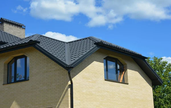 A close up on a detached house with metal roof and chimney, rain gutter system with a downspout and two windows of unusual, irregular shapes.