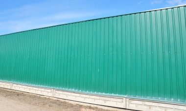 Tuğla temeli üzerine kurulmuş yeşil metal çit panelleri mavi gökyüzünü kaplıyor. 