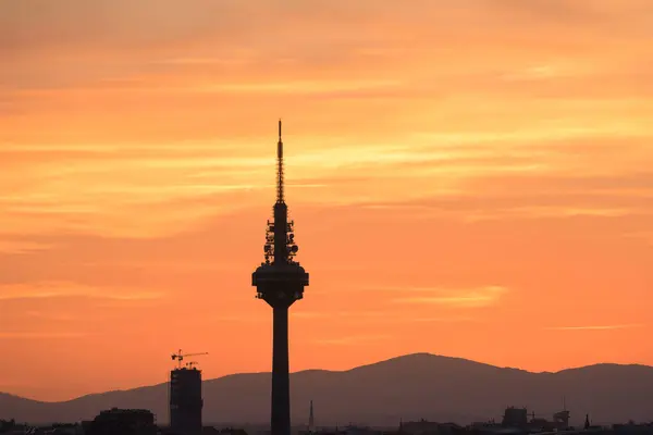 Silhouettes of buildings of a city, highlighting the silhouette of a communications tower with an orange sky with clouds at sunset. Clouds and mountains in the sky of Madrid with orange tones