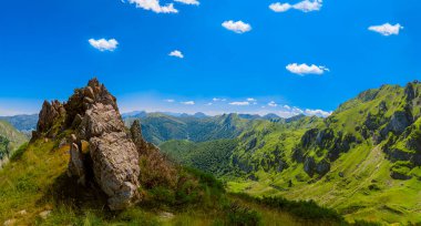 Dağ zirvesinden panoramik bir vadi manzarası. Bulutlu mavi gökyüzü. Yeşil dağ duvarları ve sonsuz gökyüzünün arkaplanı. Ön planda bir kaya