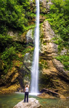Arkadan görülen kimliği belirsiz sarışın kadın, 18 metrelik bir şelaleyi işaret eden bir kayanın üzerinde duruyor ve küçük bir göle dökülüyor. Bir kaya duvarı, eğrelti otları ve yeşil dallarıyla.