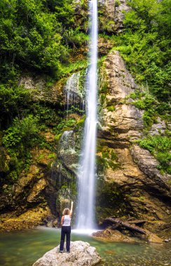 Arkadan görülen kimliği belirsiz sarışın kadın, 18 metrelik bir şelaleyi işaret eden bir kayanın üzerinde duruyor ve küçük bir göle dökülüyor. Bir kaya duvarı, eğrelti otları ve yeşil dallarıyla.