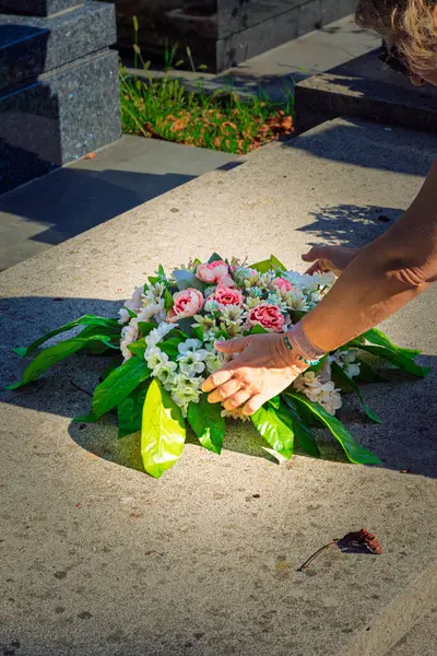Hands of an unknown woman placing a wreath of flowers on an unknown gravestone in a cemetery.