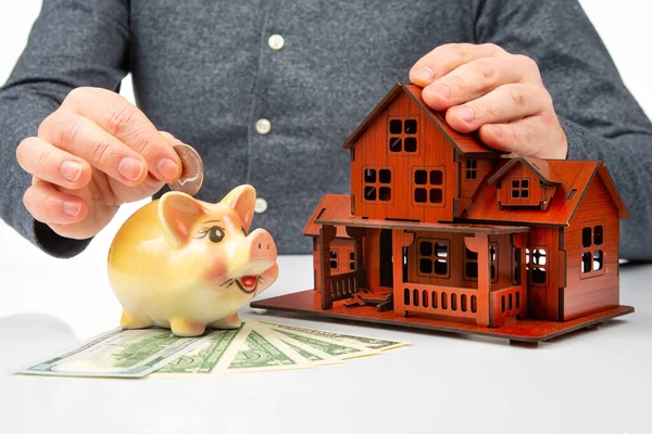 stock image piggy bank with money and a model of a house in the hands of a man. the concept of accumulating finance to buy a house